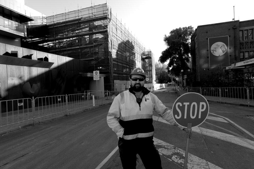 Parkville during COVID lockdown - Construction worker standing on Monash Rd with New Student Precinct construction site in background - Monochrome