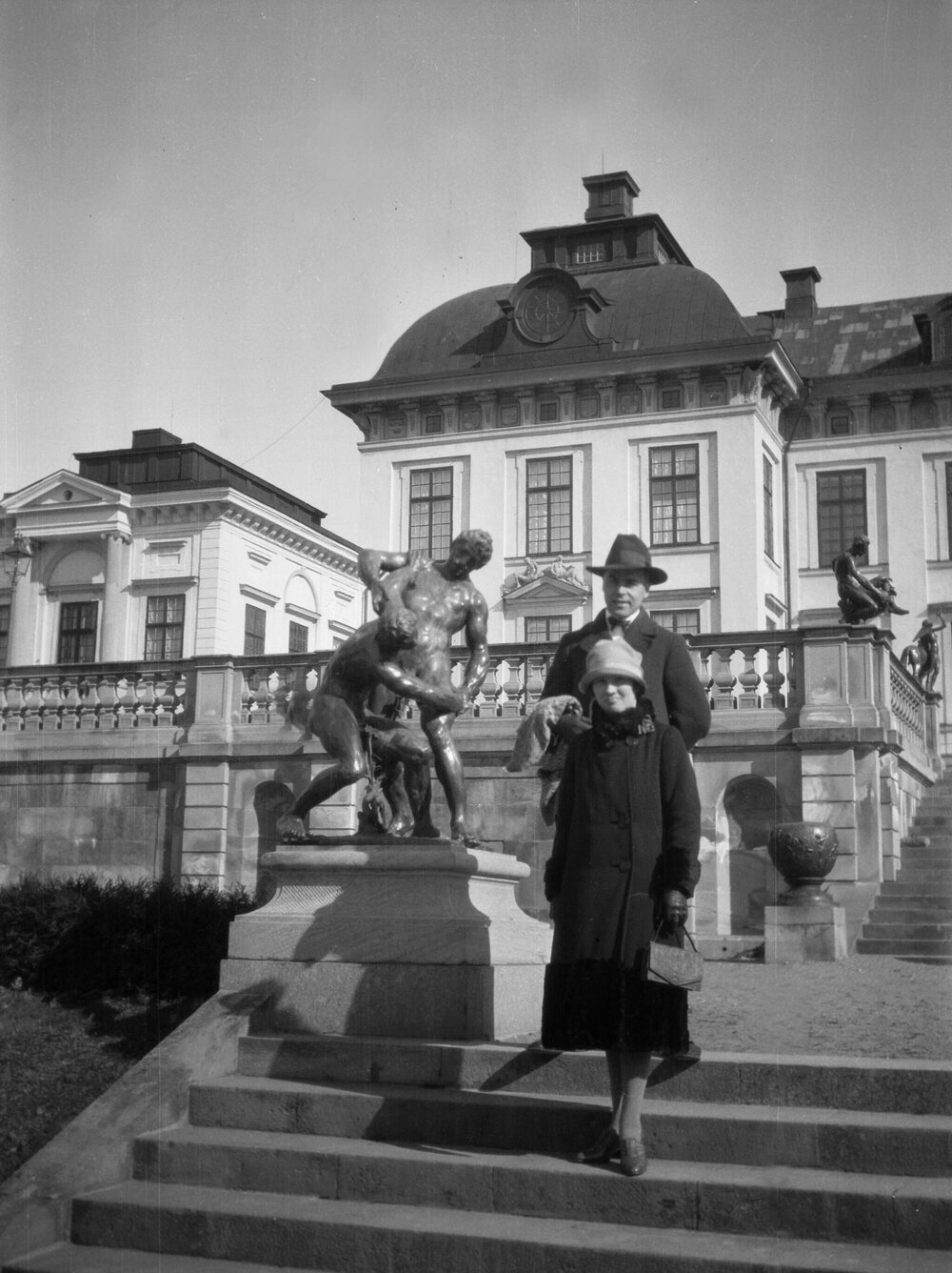 Two unidentified people alongside a statue in front of a prominent building, in an unknown Swedish city