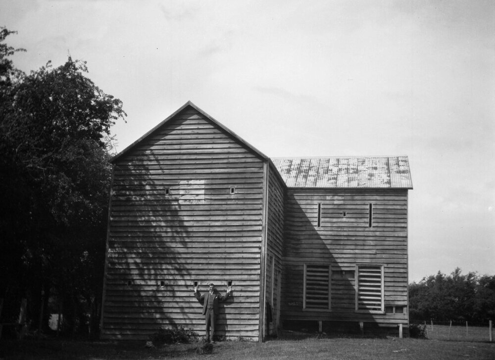 An unidentified man standing against the external wall of a large two-storied timber building in a rural setting at an unknown location in New Zealand