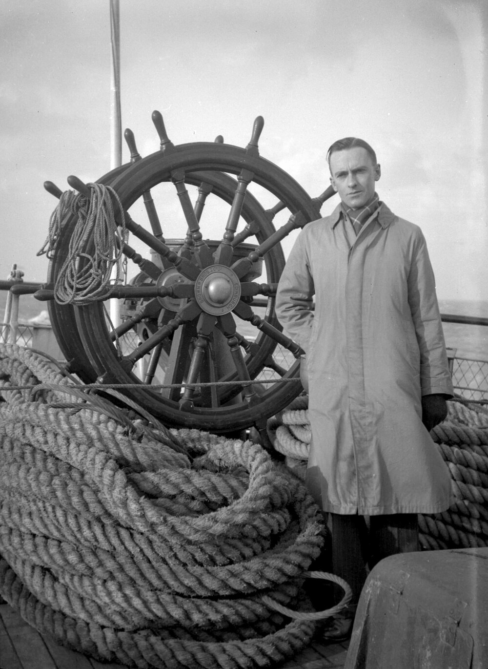 An unidentified man standing alongside a ship's wheel and coiled rope