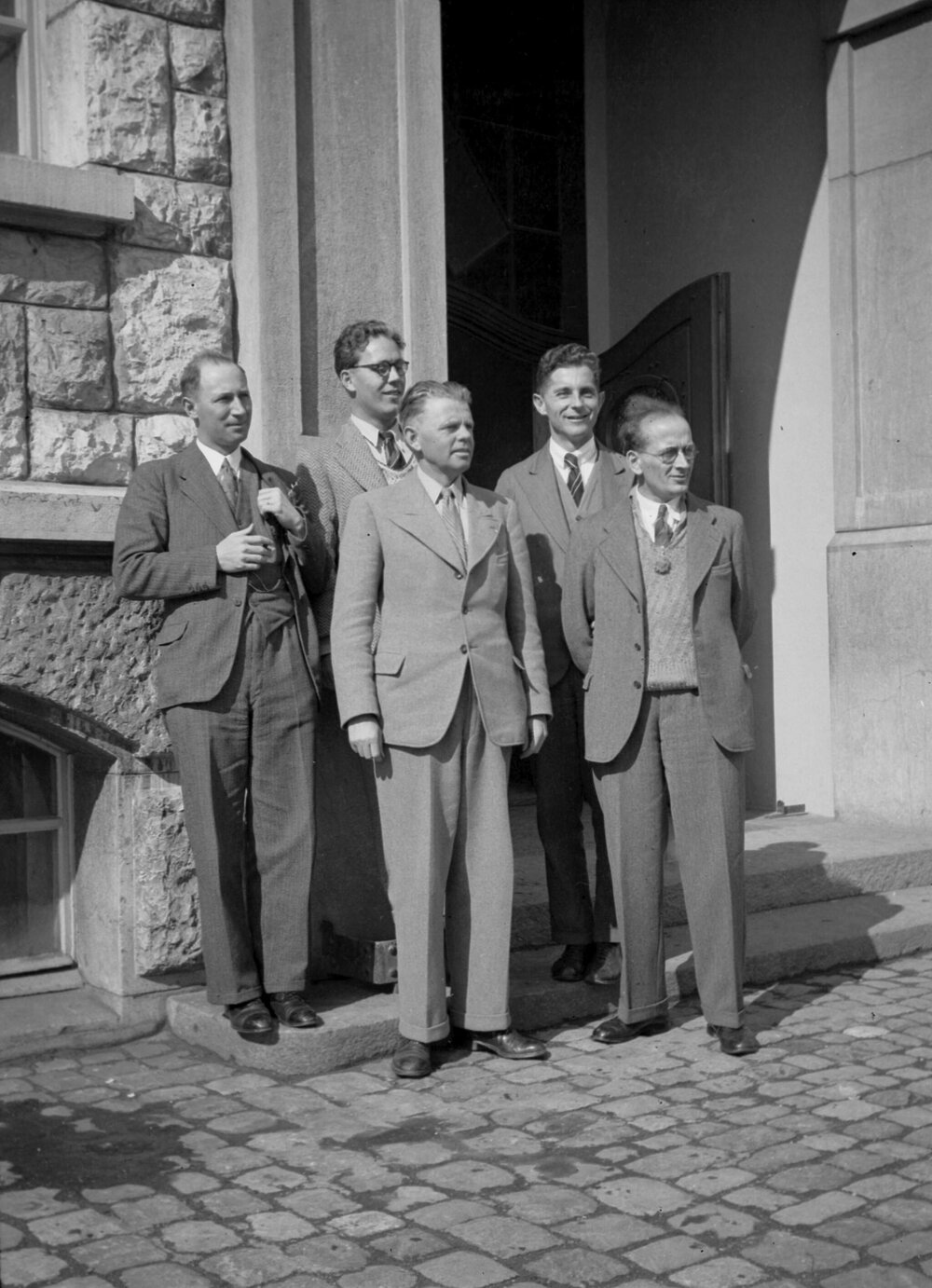 A posed group of five unidentified men dressed in suits, standing in front of a building entrance