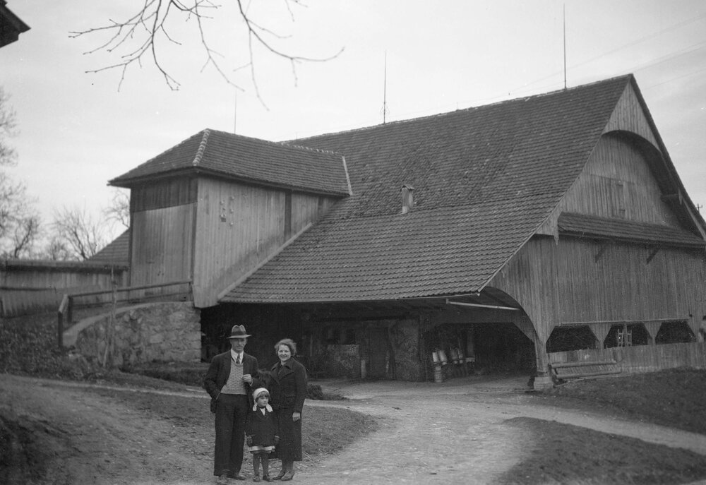 An unidentified woman, man and  small girl posed in front of a large timber, tiled building, possibly a farmhouse and dairy, at an unknown location