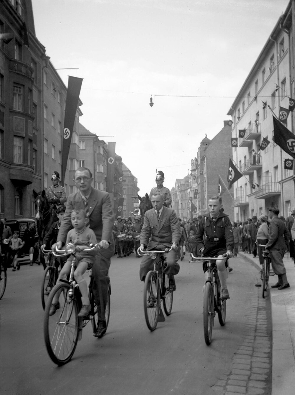 A street scene including men and boys on bicycles, parading soldiers and Nazi flags