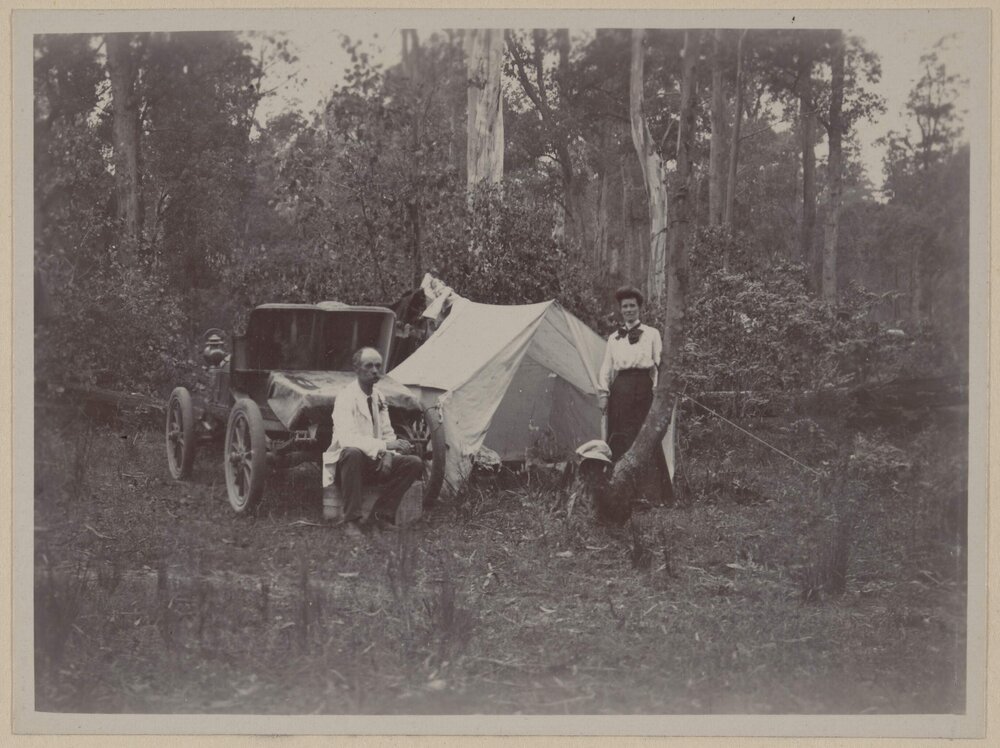 [Seated man and woman standing next to tent and motor car]