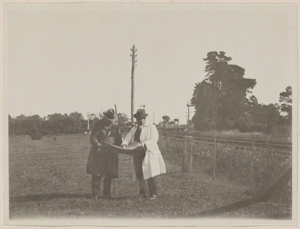 [Two men viewing document, standing next to railway line]