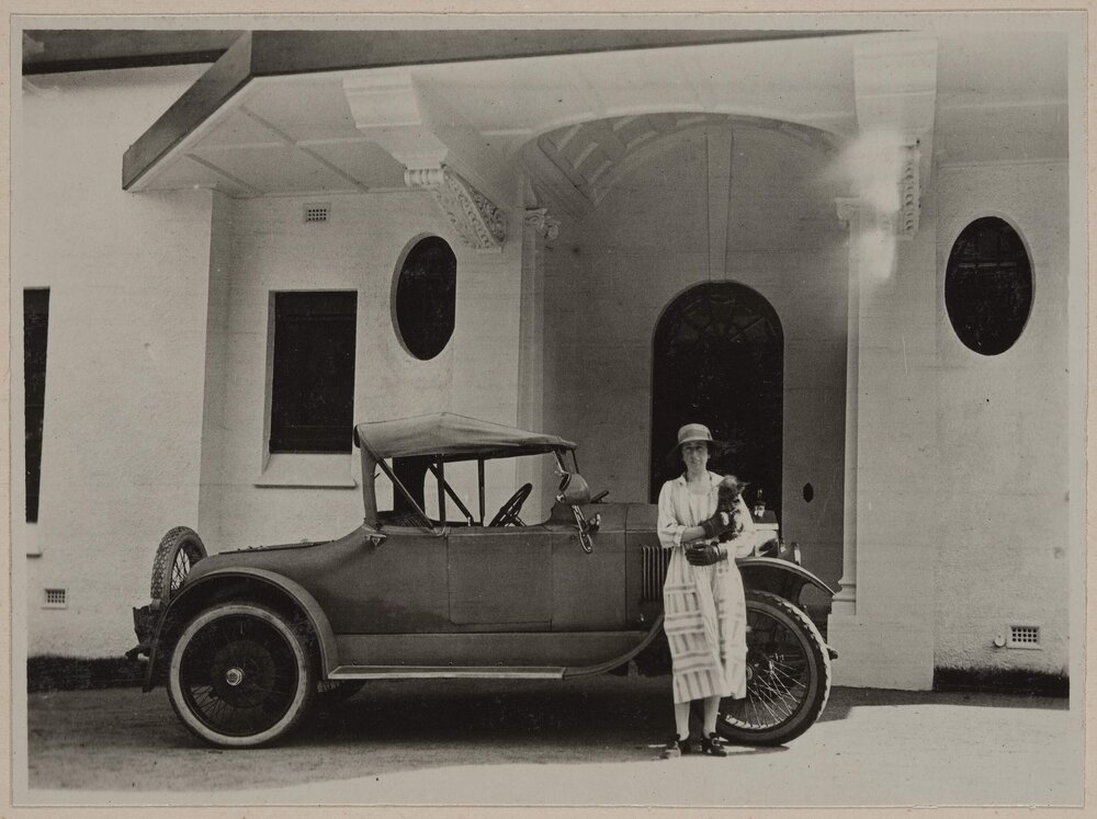 [Woman holding terrier dog posing next to motor car]