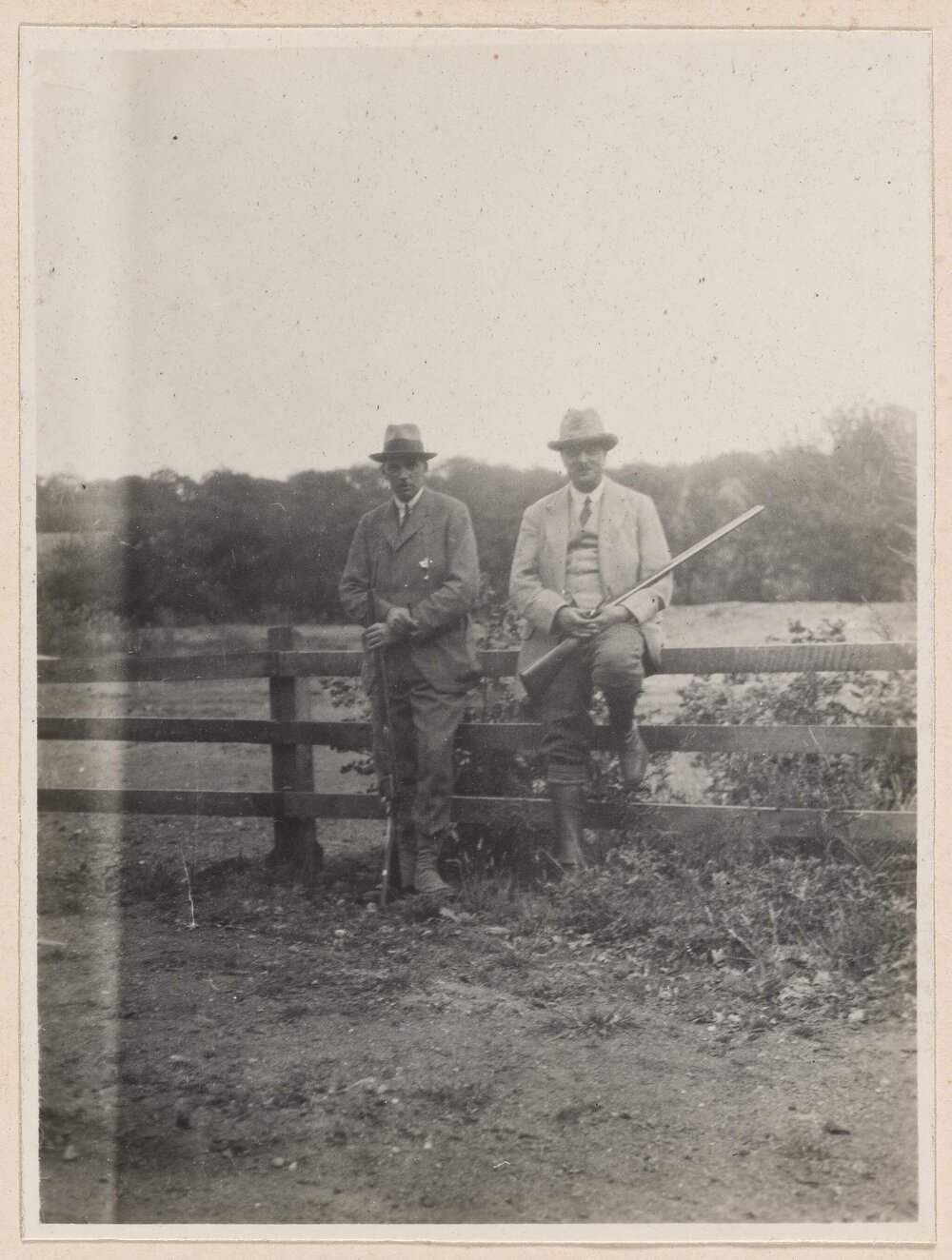 [Two men holding shotguns posing on fence]