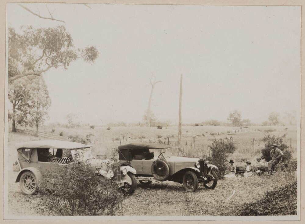 [Man and two women sitting next to two motor cars]