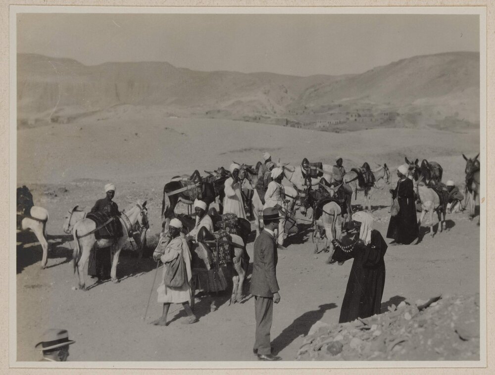 [Man wearing suit and hat standing in front of group of men wearing robes and turbans]