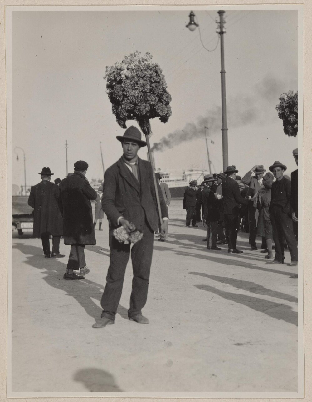 [Man wearing suit and hat holding arrangement of flowers on long wooden stick]