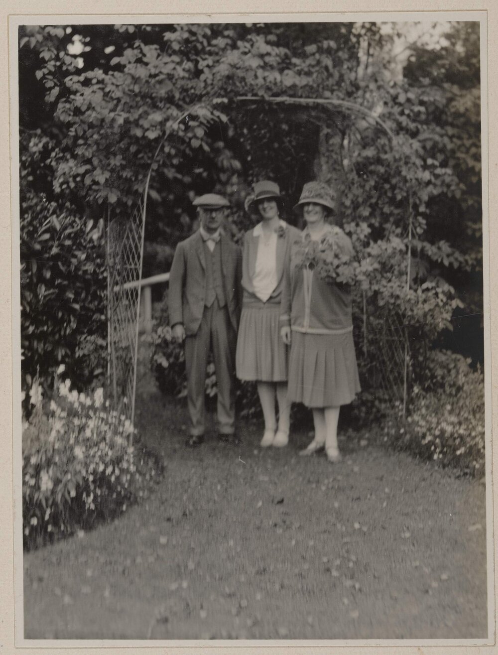 [Man and two women standing under garden arch trellis]