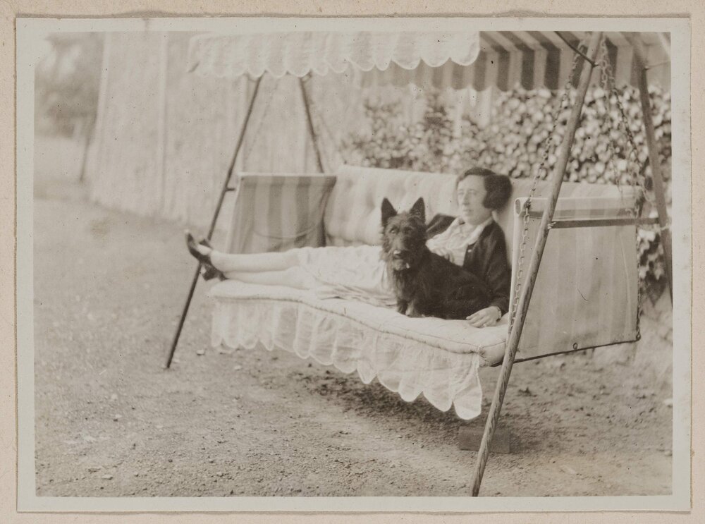 [Woman and terrier dog posed seated on  outdoor swing chair]