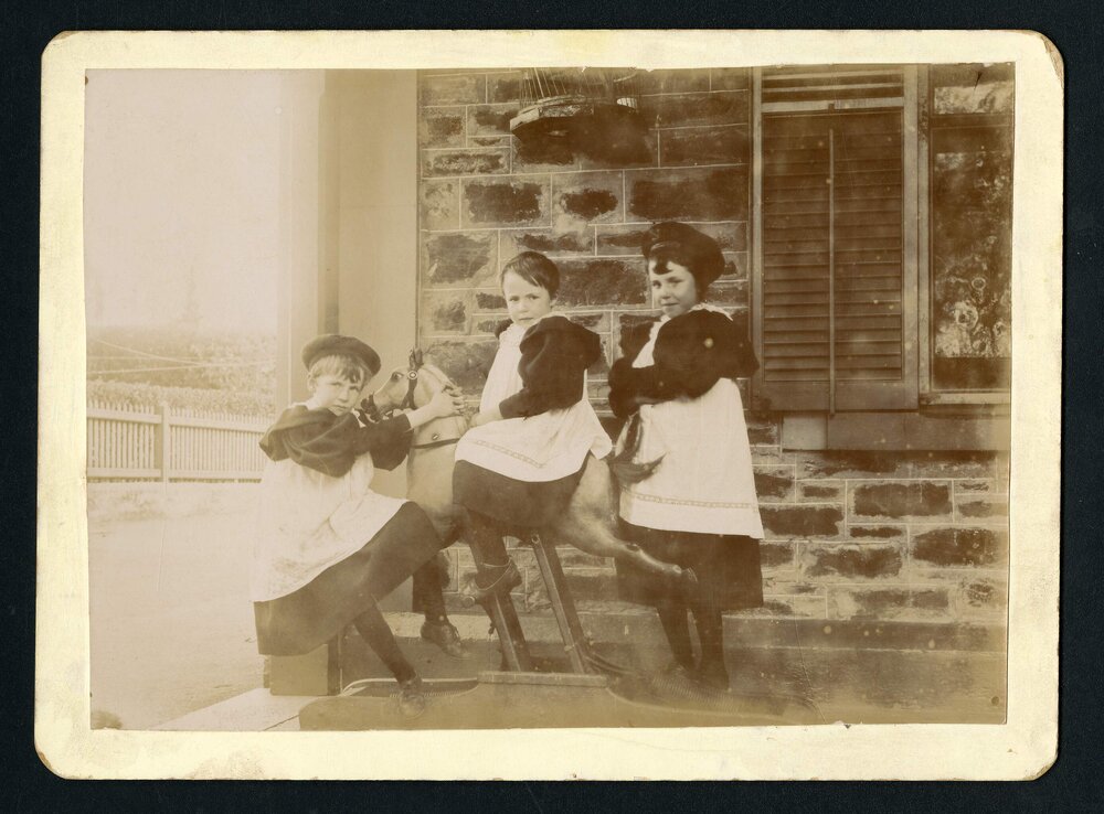 Three girls on rocking horse on verandah of house
