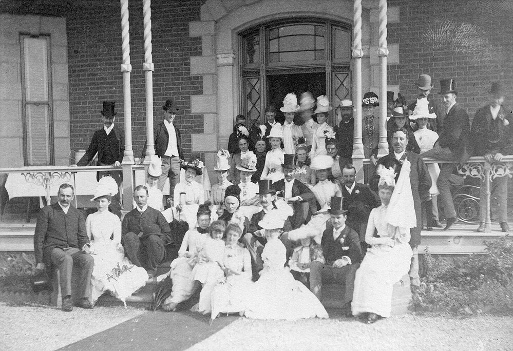 Large group, men and women, seated on front steps of house