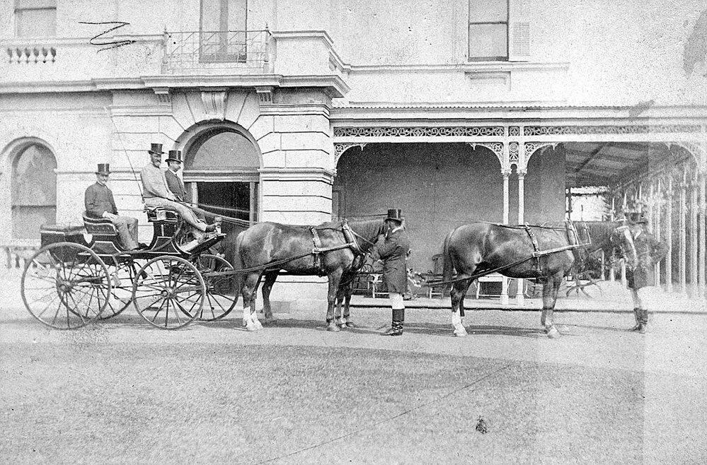 Photo of three men in horse drawn carriage, grooms attending horses