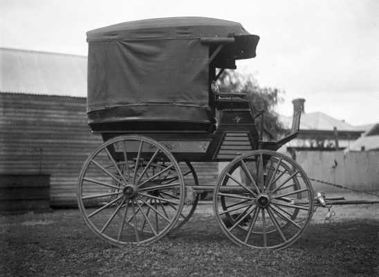 A covered wagon or wagonette outside a timber building.