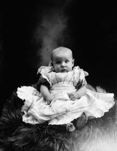 Studio portrait of a baby wearing a lace dress.   Family name Thurston.