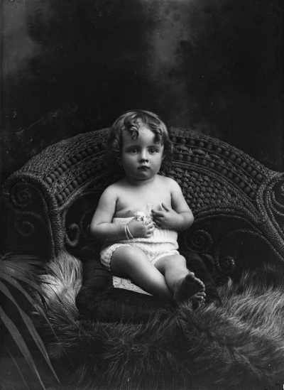 Studio portrait of a young child seated in a large wicker chair.  Family name Condon.