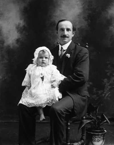 Studio portrait of a seated man holding a bare-footed baby.  Family name Walsh.