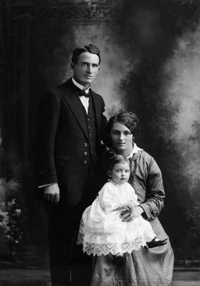 Studio portrait of woman holding a baby and seated alongside a man.  Family name Holmes.