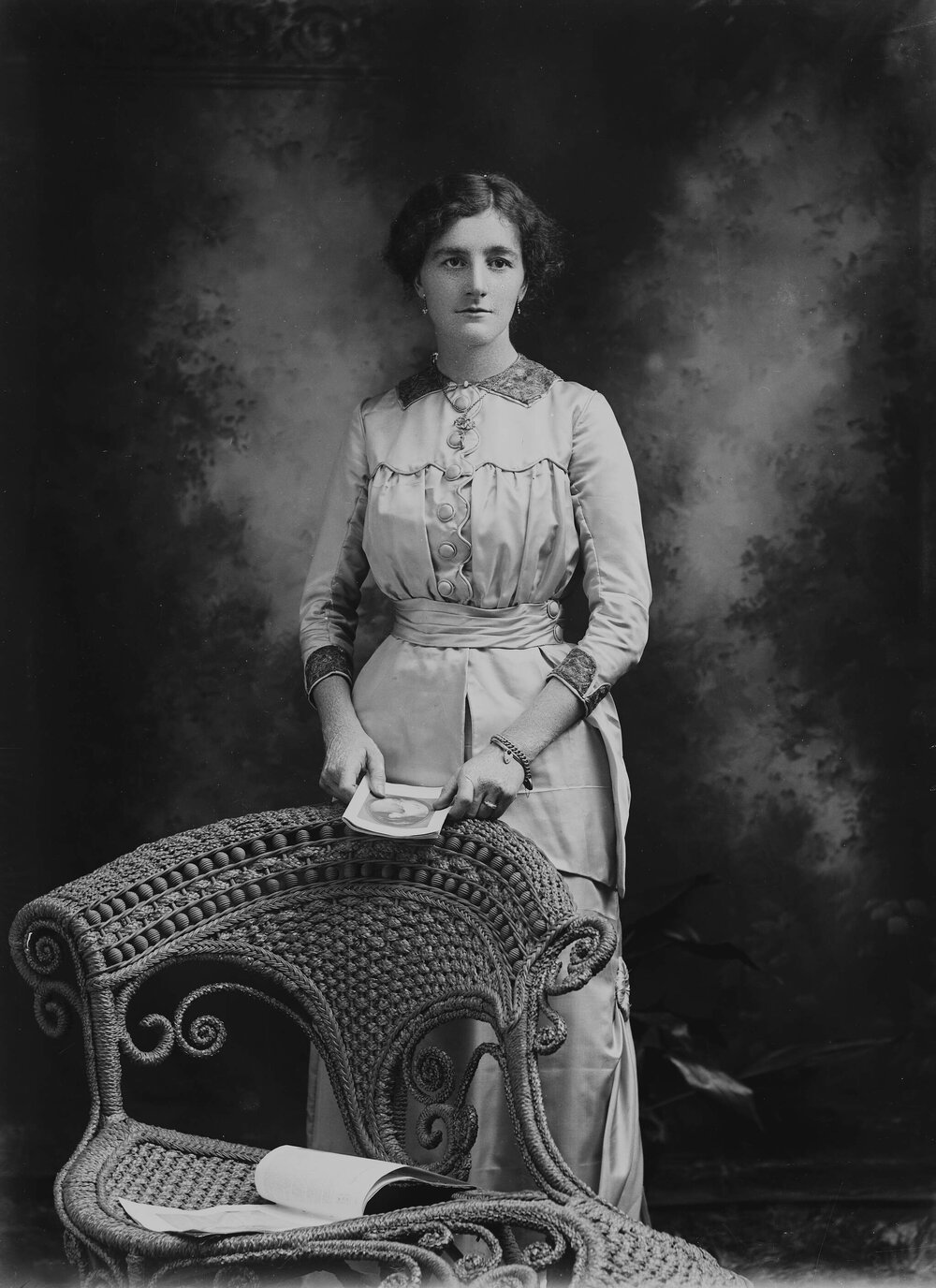 Studio portrait of a woman standing in front of a wicker chair.  Possible name Miss Stebbing.