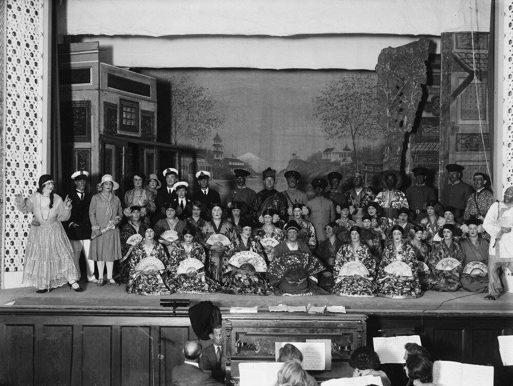 A large group of performers on a stage, some dressed as geishas.