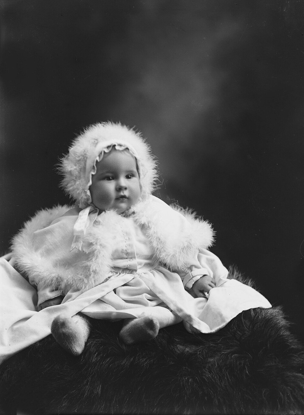Studio portrait of a baby wearing a fur trimmed bonnet and cape.  Family name Rowell or Howell.