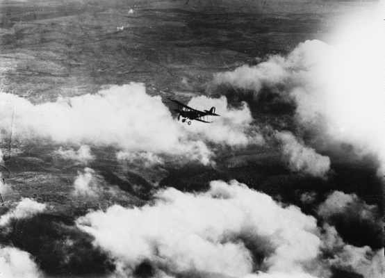 An aerial view of a single engine biplane flying above cloud and unidentified countryside.