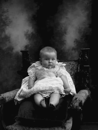Studio portrait of a baby wearing a lace dress.   Family name McNamara.