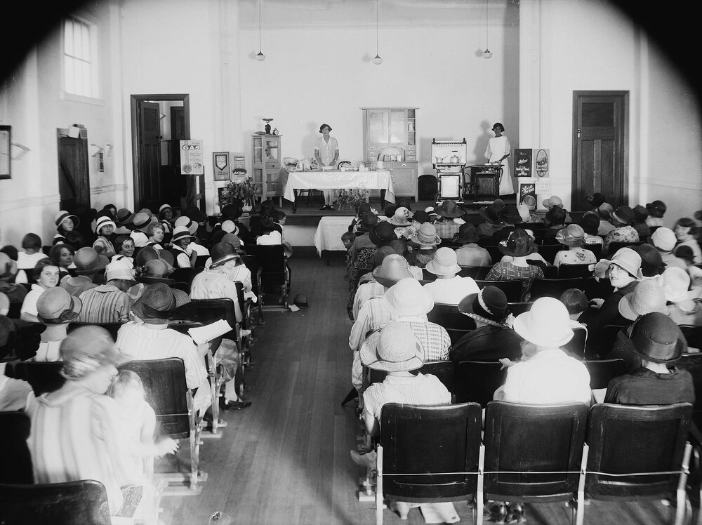 A gas appliance and cooking demonstration in a hall filled with women.