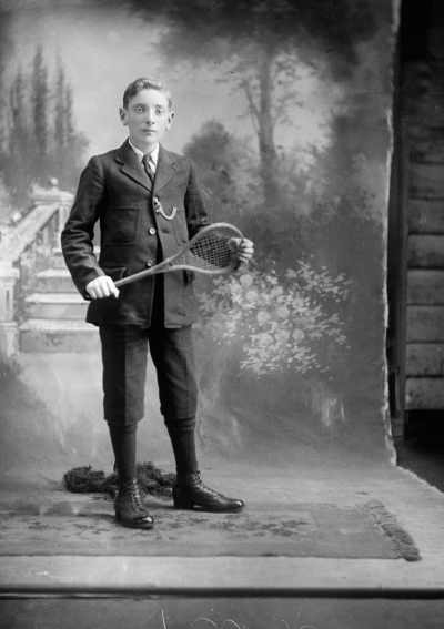 Studio portrait of a young suited man holding a stringed racquet.  Family name Corrigan.