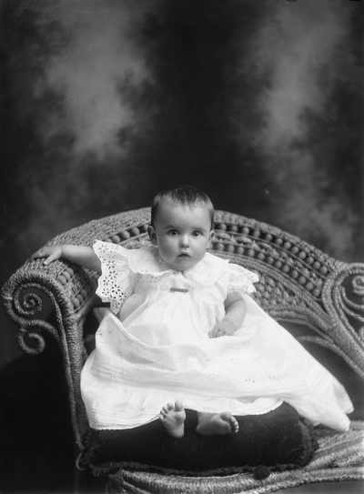 Studio portrait of a baby seated on a cushion. Family name Speedie.