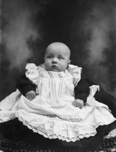 A studio portrait of a baby seated on a cushion.  Family name Rawlings.