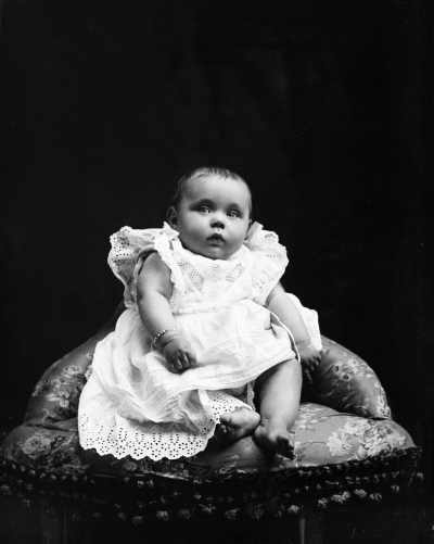 Studio portrait of a baby wearing a dress and seated on a corner chair.