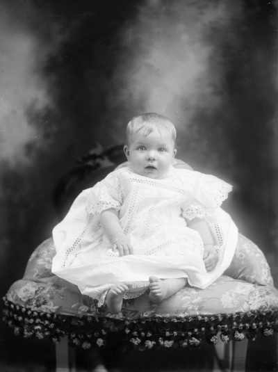 Studio portrait of a baby seated on an upholstered corner chair. Family name West.