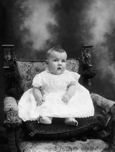 Studio portrait of a baby seated on a cushion and a carved chair.  Family name Carroll.