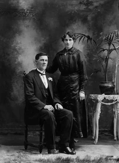 Studio portrait of a  woman wearing a black dress and a man wearing a suit and bow tie.  Family name Trethowan.
