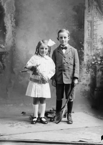 Studio portrait of a boy holding a stringed racquet and girl holding a large lily.   Family name Taylor.