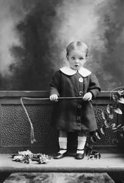 Studio portrait of an infant holding a riding crop.  Family name Petersen.