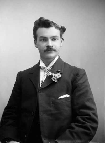 Portrait of a young man wearing a sprig of flowers in his button hole.