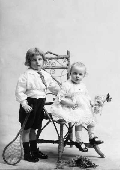 Studio portrait of  a young boy standing alongside a seated baby girl .   Family name Dynon.