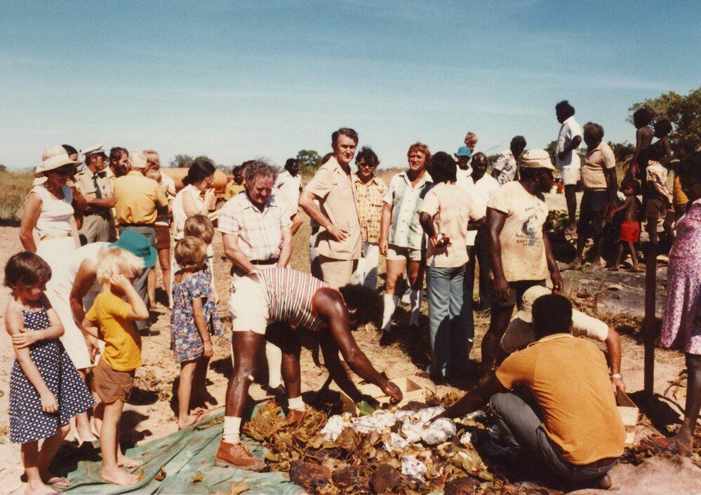 Malcolm Fraser in the Northern Territory standing with others gathered around food being cooked in an earth oven.