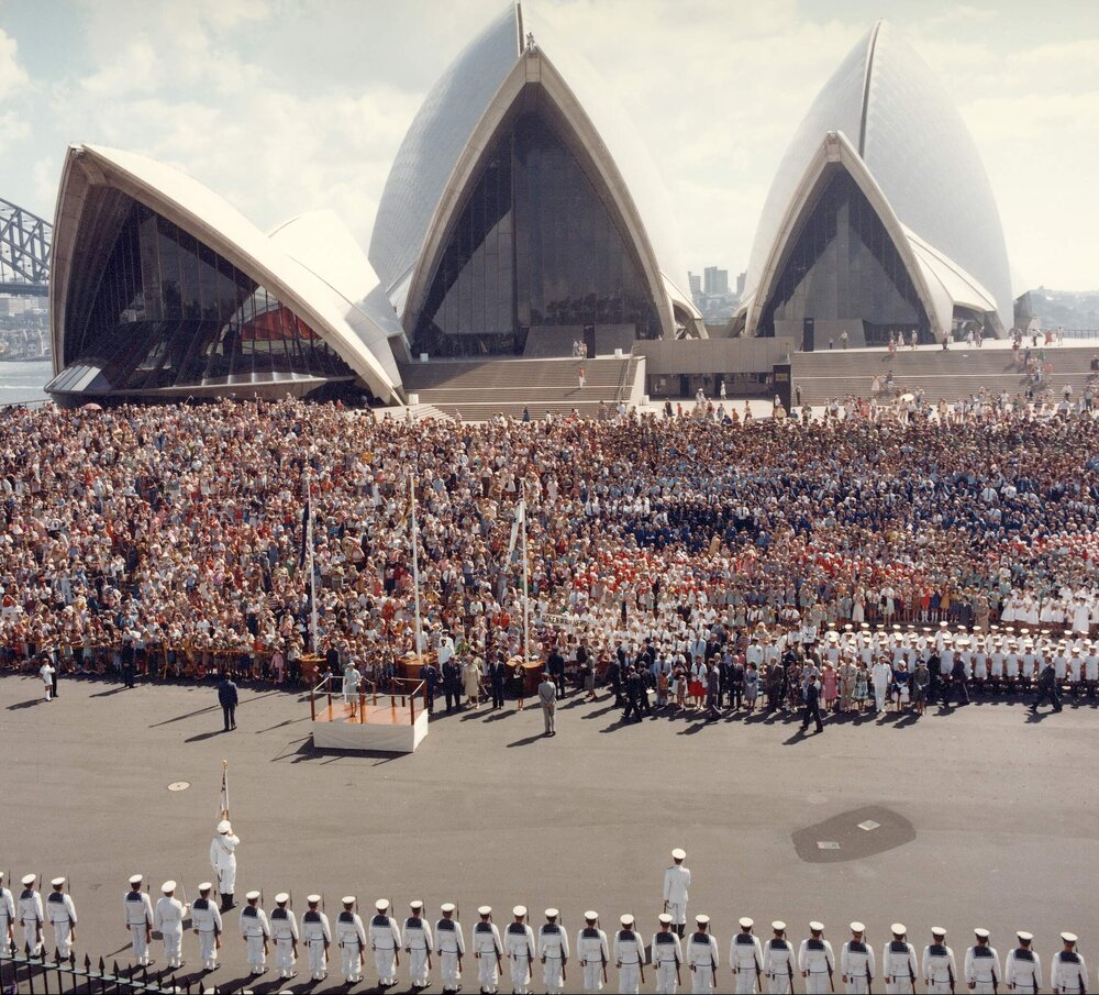 Queen Elizabeth II standing on a dais on the forecourt of the Sydney Opera House after her arrival in Sydney as part of her 1977 official Australian visit.
