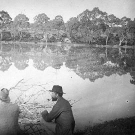 Two unidentified men squatting by an unidentified lake,