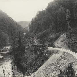 A view of a locomotive crossing a small timber trestle railway bridge alongside the King River, Tasmania.  Part of the operations of the Mount Lyell Mining & Railway Co. Ltd.