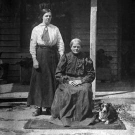 Two women and a dog in front of a timber dwelling.