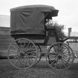 A covered wagon or wagonette outside a timber building.