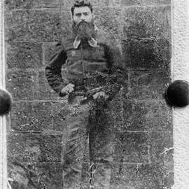 Ned Kelly, shackled and standing against a stone wall.