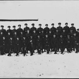 A large unidentified group of men in military uniforms posed against building in snow.