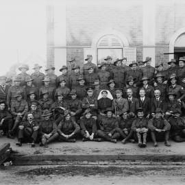 A large group including men in military uniform and one nurse, posed outside a brick building.